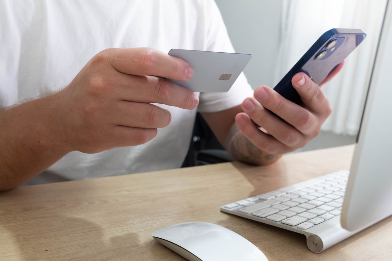 services-02 Person holding credit card and smartphone for an online transaction at a desk.